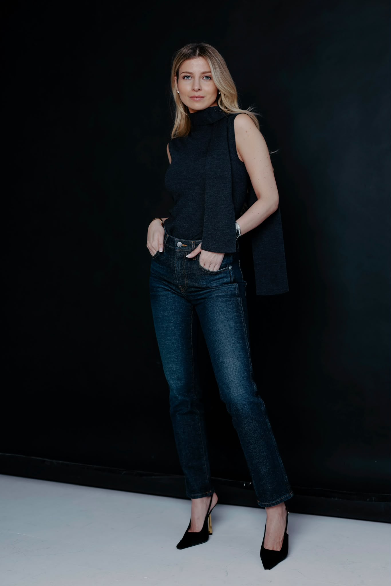 Agne Skopaite, standing, in a black knit and dark denim against a black studio backdrop